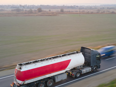 An 18-wheeler towing a tank of hazardous materials drives fast on a highway.
