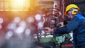 Factory worker in a yellow hard hat, safety glasses, and ear protection operating heavy industrial machinery in a manufacturing plant