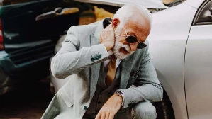 An older man in a suit crouches in front of a wrecked vehicle, holding his neck — a possible indication of whiplash, one of the most common car accident injuries.