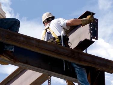 Two ironworkers in hard hats and safety harnesses perched on steel I-beams high above the ground during structural construction, illustrating dangerous elevation work