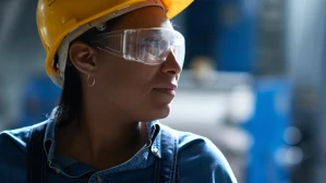 Female industrial worker wearing a yellow hard hat and safety glasses looking off to the side in a factory setting, representing workers