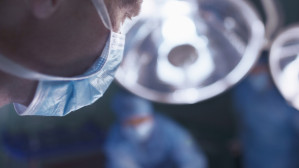 Close-up profile of a masked surgeon under bright OR lights with surgical team visible behind, representing medical malpractice and physician negligence