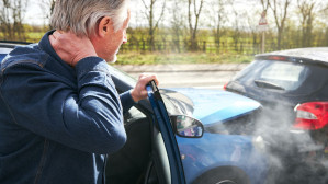 Older man clutching his neck in pain after a rear-end car accident, steam rising from vehicle