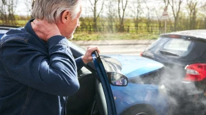 Older man clutching his neck in pain after a rear-end car accident, steam rising from vehicle