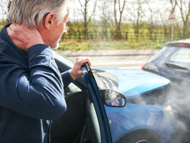 Older man clutching his neck in pain after a rear-end car accident, steam rising from vehicle
