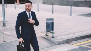 A pedestrian accident lawyer with a beard and glasses in a black suit crosses the street in a crosswalk while carrying a black briefcase.