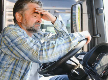 A man, with his eyes closed, holds on to a truck’s steering wheel and tries to shake off truck driver fatigue.