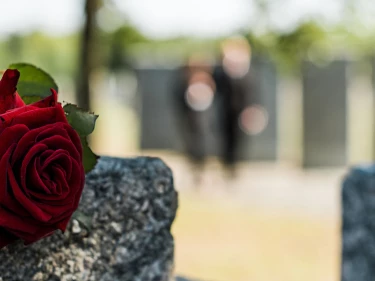 Red rose resting on a granite headstone in a cemetery with mourners in the background, representing the grief and loss that leads families to pursue a wrongful death lawsuit